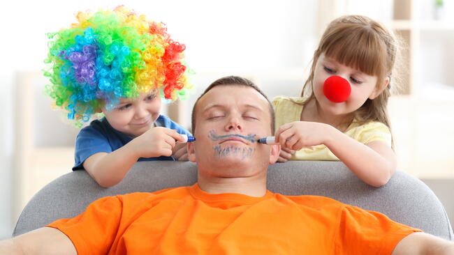 Two kids dressed as clowns draw on their sleeping father's face with markers