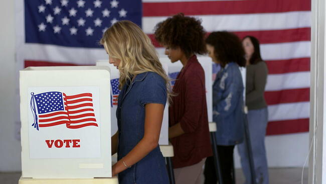 Four women of various demographics, young blonde woman in front, filling in ballots and casting votes in booths at polling station, US flag on wall at back. Focus on booth signage